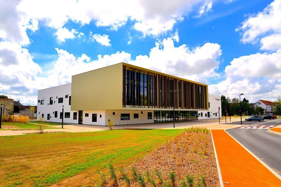 Un bâtiment moderne de deux étages avec une façade à dominante blanche et des pans de jaune et de grandes baies vitrées. Il est entouré d'un espace vert paysager, d'un chemin rouge et d'un ciel bleu clair parsemé de nuages.