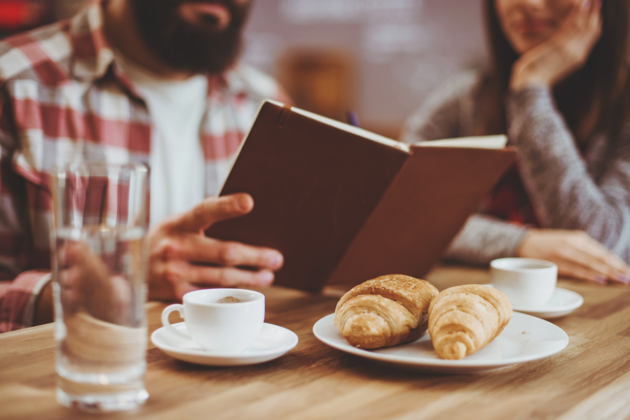 Une personne lit un livre sur une table en bois dressée avec deux croissants dans une assiette, deux tasses de café et un verre d'eau. Une autre personne est assise à proximité, partiellement visible. La scène suggère une atmosphère détendue et chaleureuse.