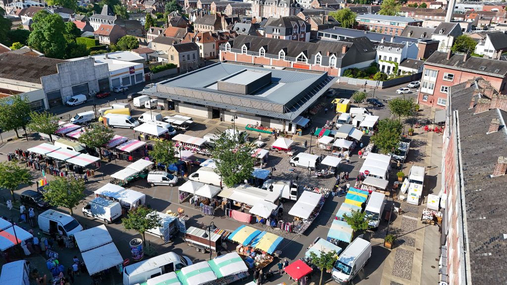Vue aérienne d'un marché en plein air animé avec de nombreux stands et des auvents colorés. Les gens font leurs courses et se promènent. Autour du marché se trouvent divers bâtiments et des rues bordées d'arbres dans un cadre urbain.