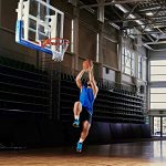 Un joueur de basket-ball en maillot bleu et short noir saute vers le panier pour un dunk sur un terrain couvert. Des lumières vives au plafond illuminent le plancher en bois et les gradins vides en arrière-plan.