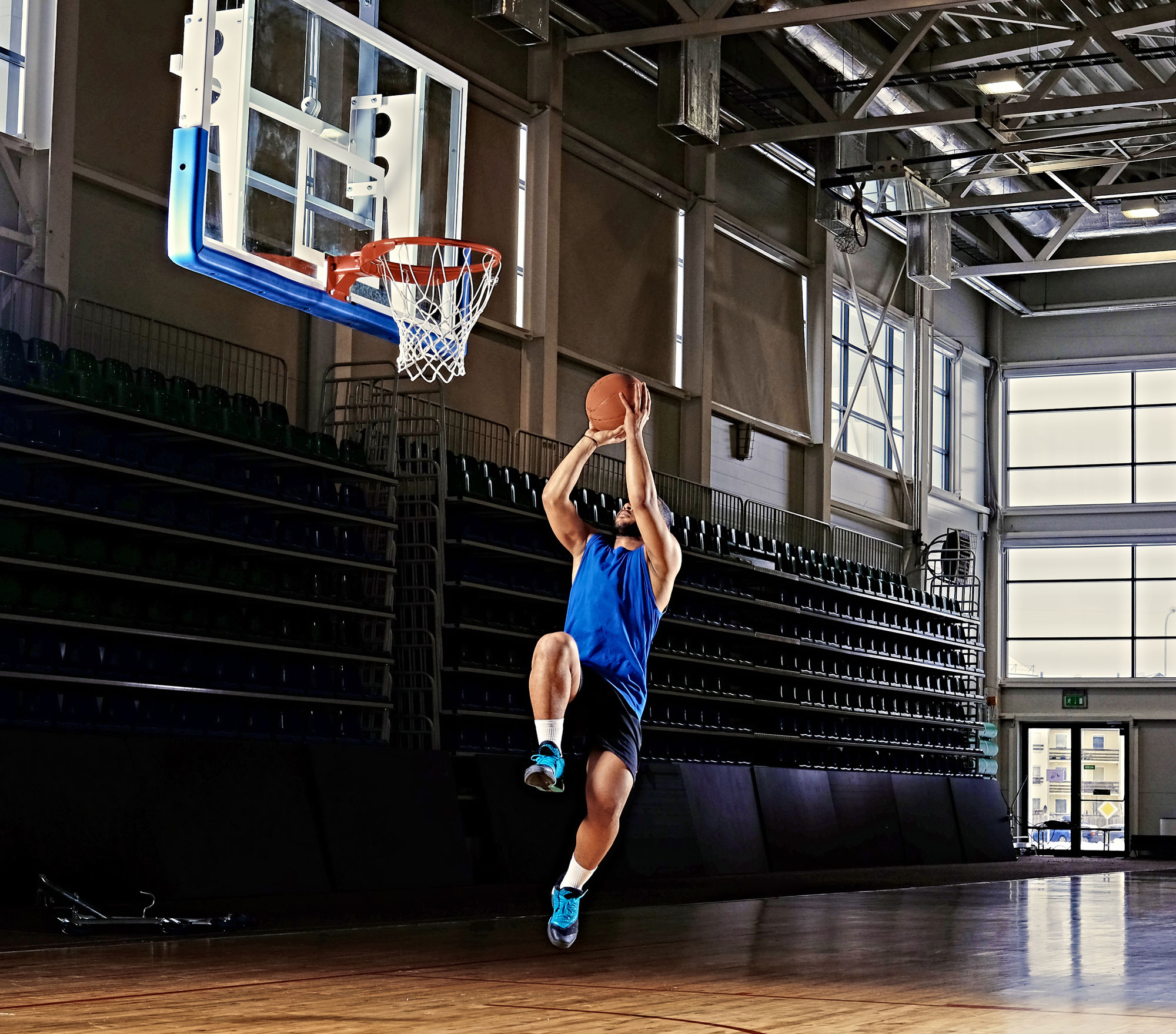Un joueur de basket-ball en maillot bleu et short noir saute vers le panier pour un dunk sur un terrain couvert. Des lumières vives au plafond illuminent le plancher en bois et les gradins vides en arrière-plan.