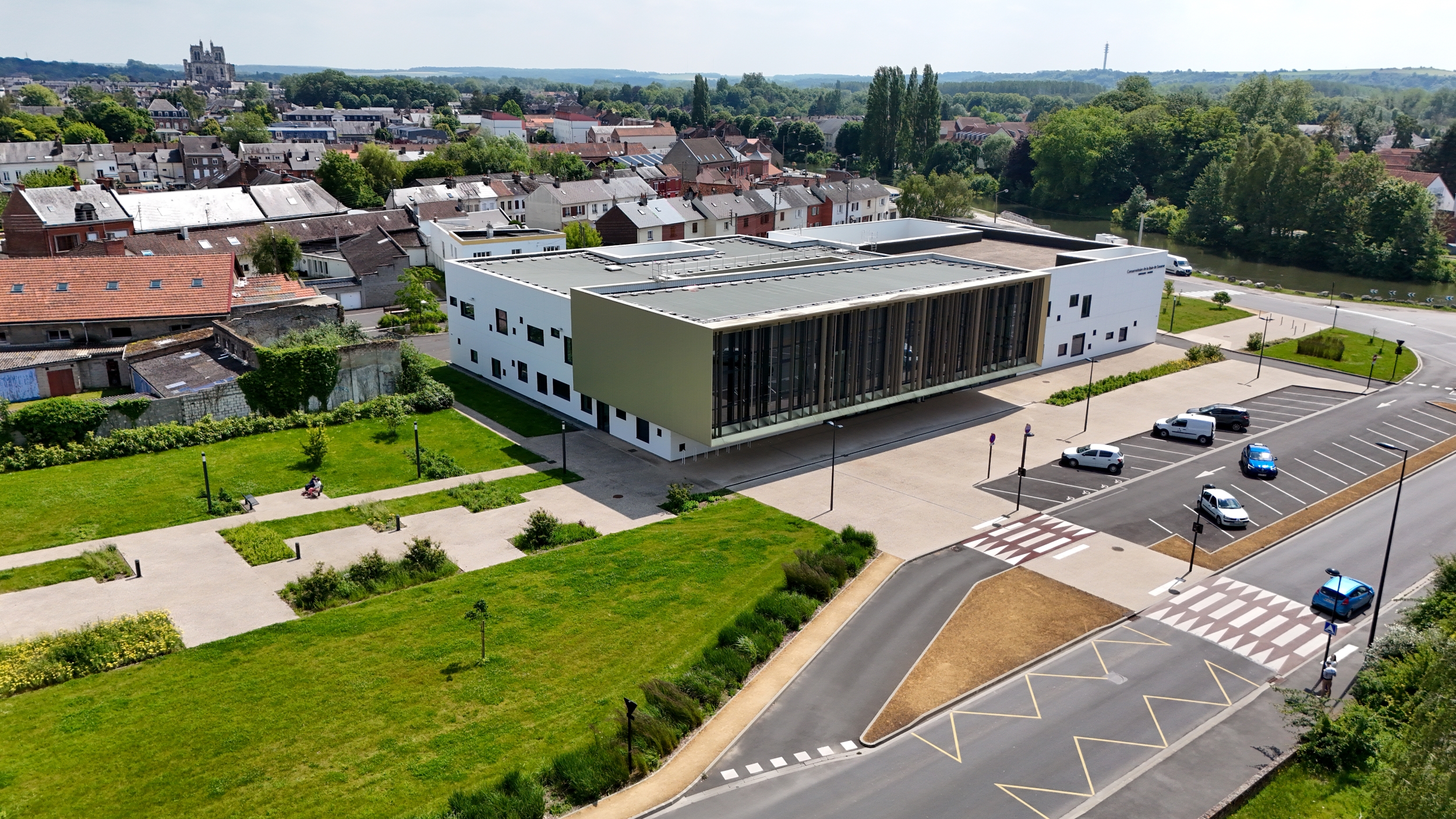 Vue aérienne d'un bâtiment moderne rectangulaire avec une façade en verre, entouré de verdure et de sentiers. Le bâtiment est situé dans une zone suburbaine avec des maisons résidentielles à proximité et une route avec quelques véhicules garés au premier plan.